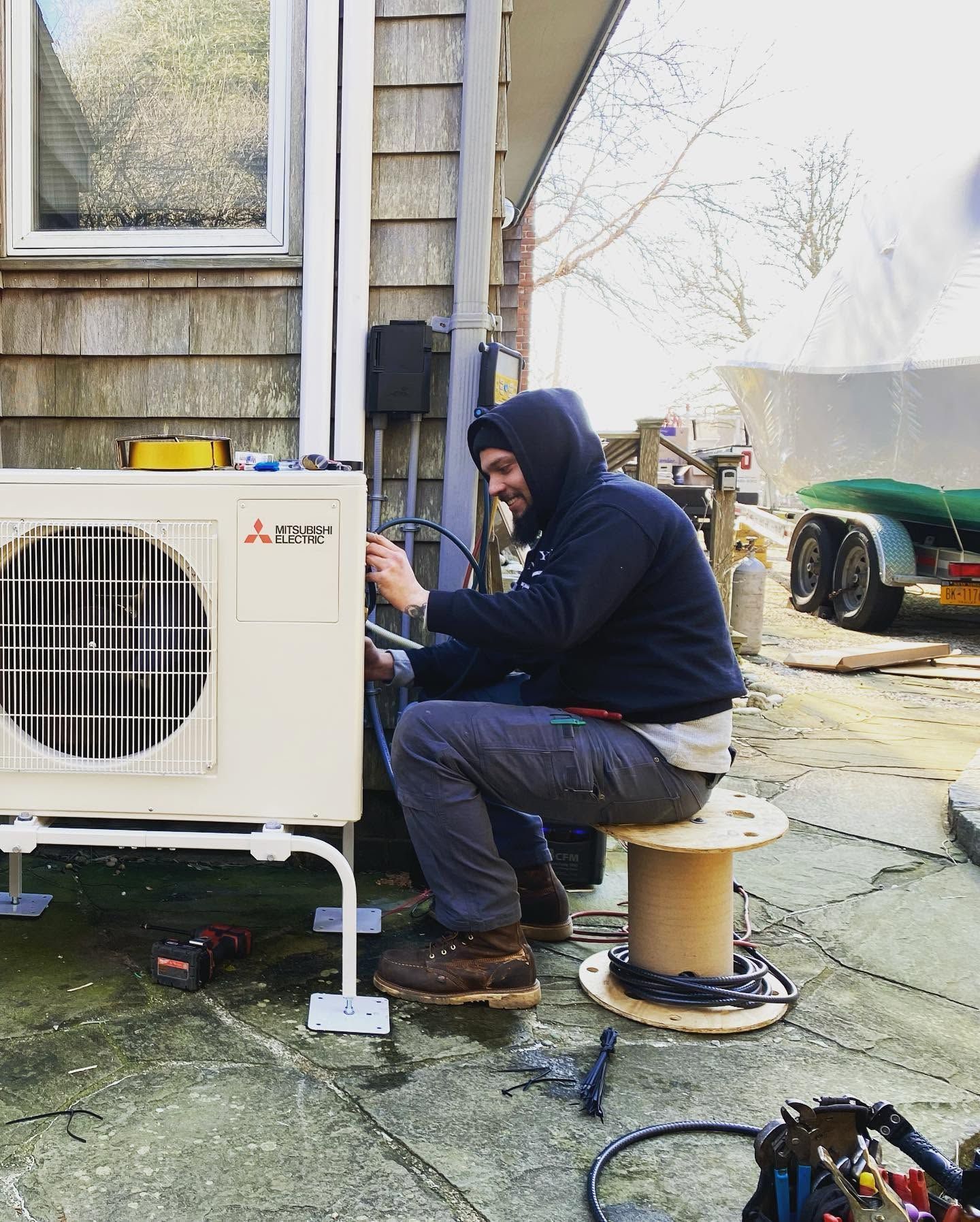 Technician kneels beside an outdoor HVAC unit at a house, inspecting or repairing it.