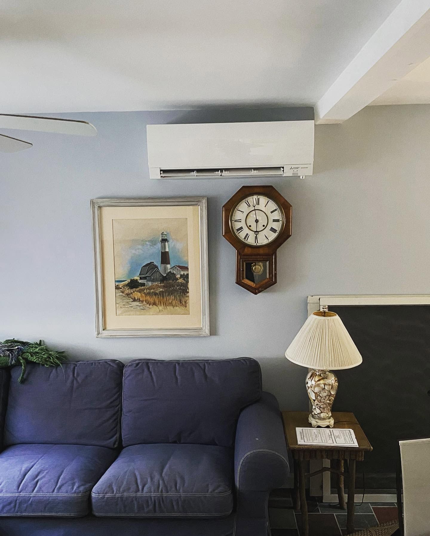 Cozy living room with blue sofa, wall clock, framed painting, table lamp, and wall-mounted air conditioner.