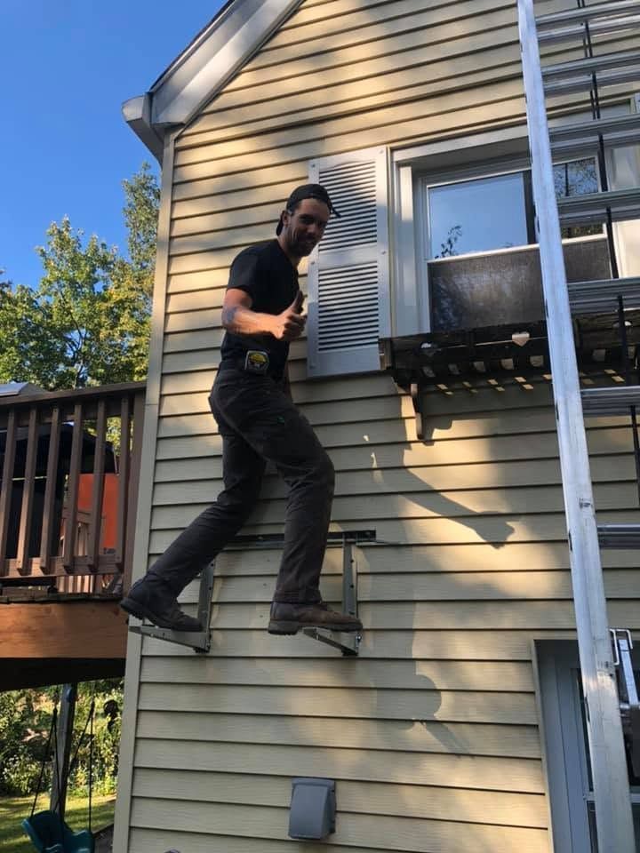 Person climbing a house wall near a window and deck, wearing black clothes and safety boots