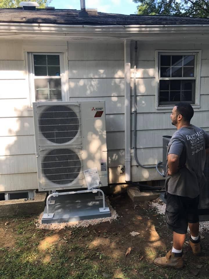 Outdoor HVAC unit beside a house; a man stands nearby inspecting it.