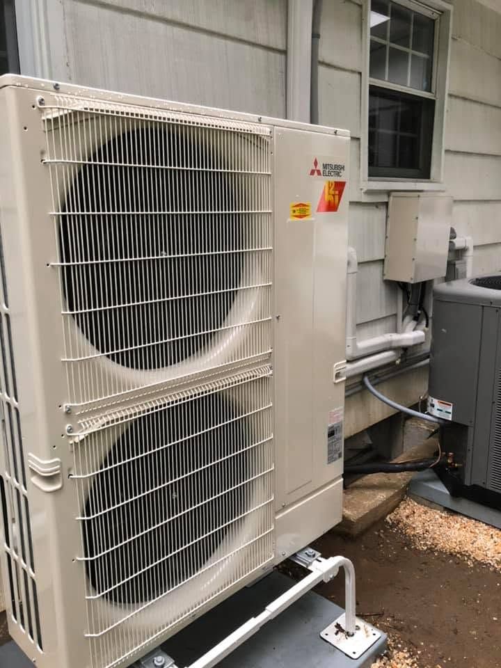 Outdoor HVAC condenser unit beside a building wall with pipes and vents, white housing and metal grille.