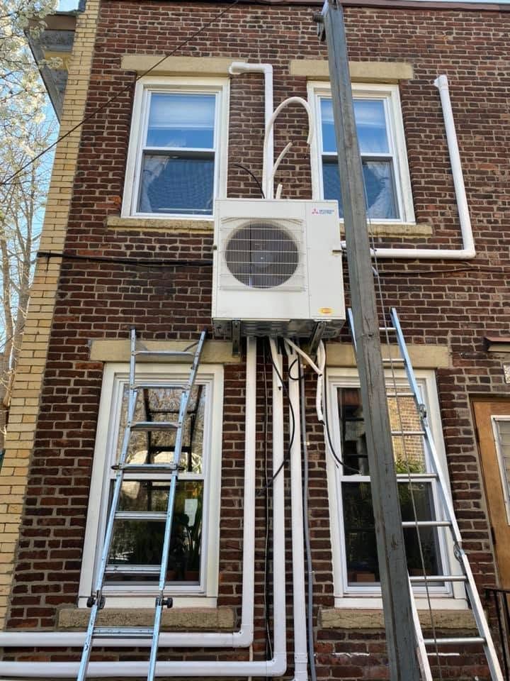 Brick building facade with windows, exterior AC unit, and several pipes and ladders attached to the wall.