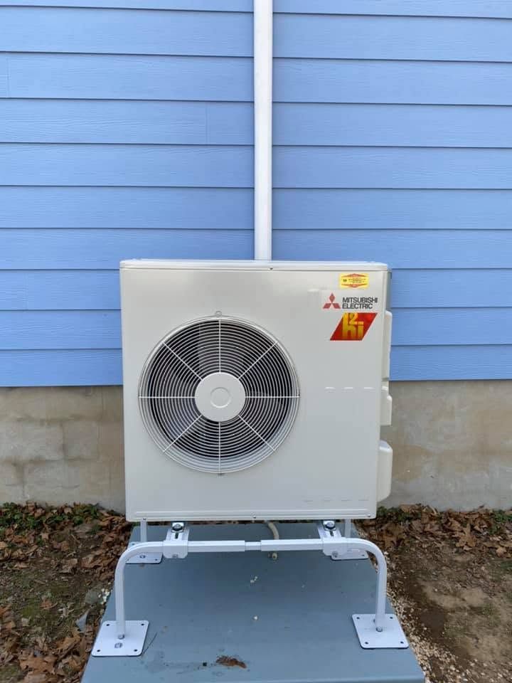 Outdoor wall-mounted air conditioner unit against a light blue exterior wall, with a metal support frame on concrete.