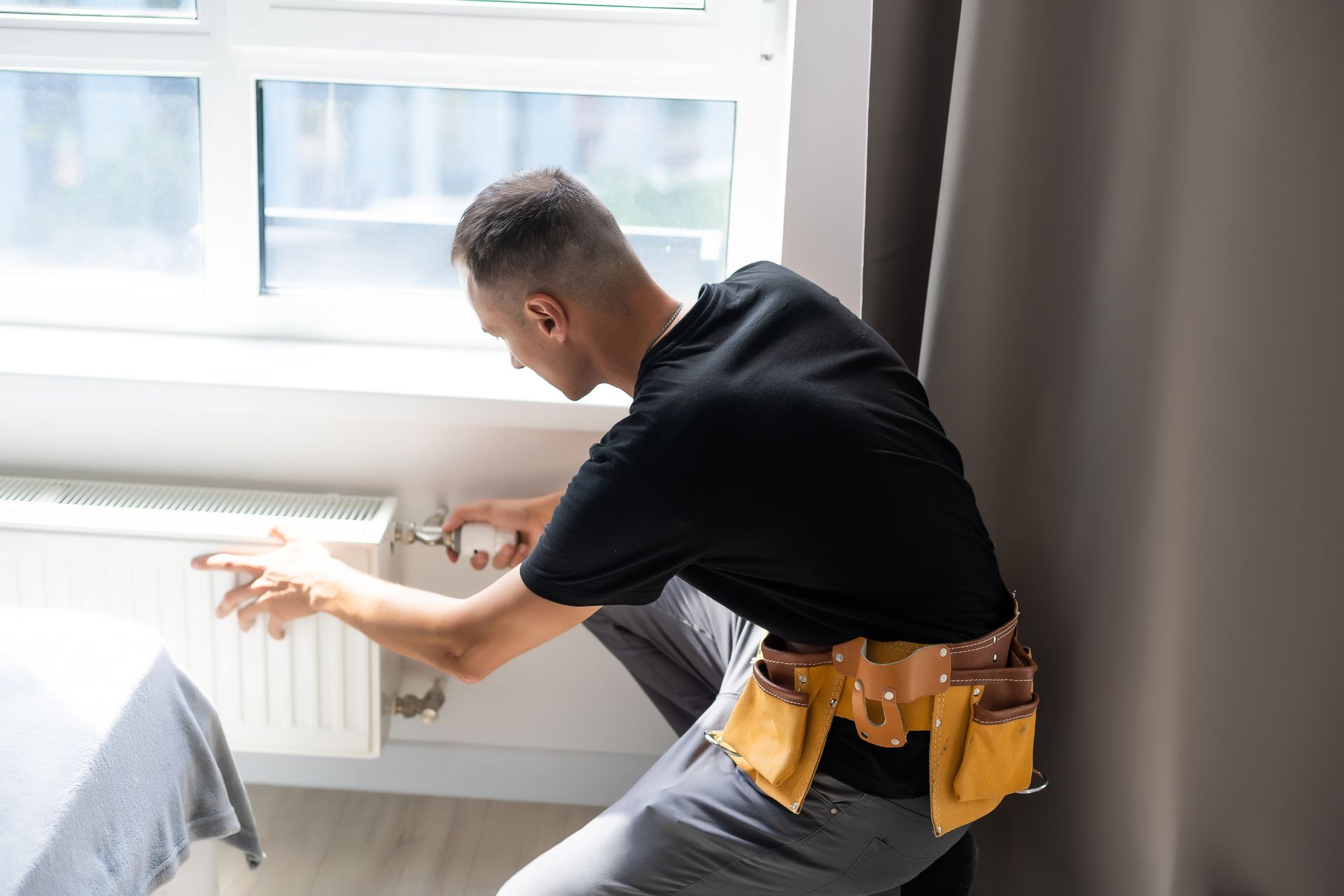 Technician adjusting a home radiator beneath a window while performing maintenance.