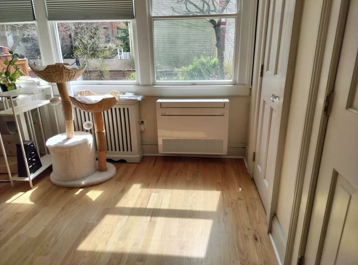 Bright sunlit room with wooden floor, window, radiator, white cat tree, and door on the right.