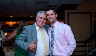 Two men smiling and posing together indoors, one in a green suit giving a thumbs-up
