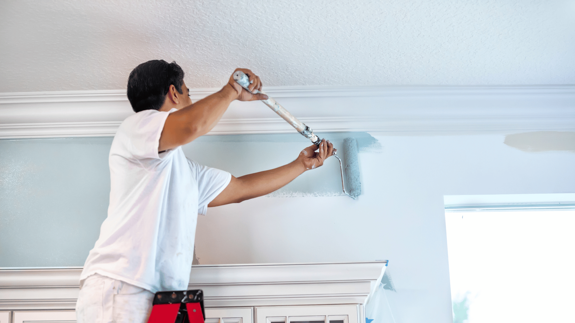 A man is painting the ceiling of a room with a paint roller.