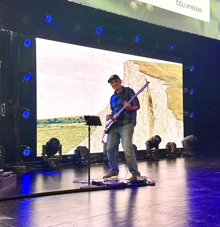 Musician playing electric guitar onstage under blue lights with a large screen backdrop