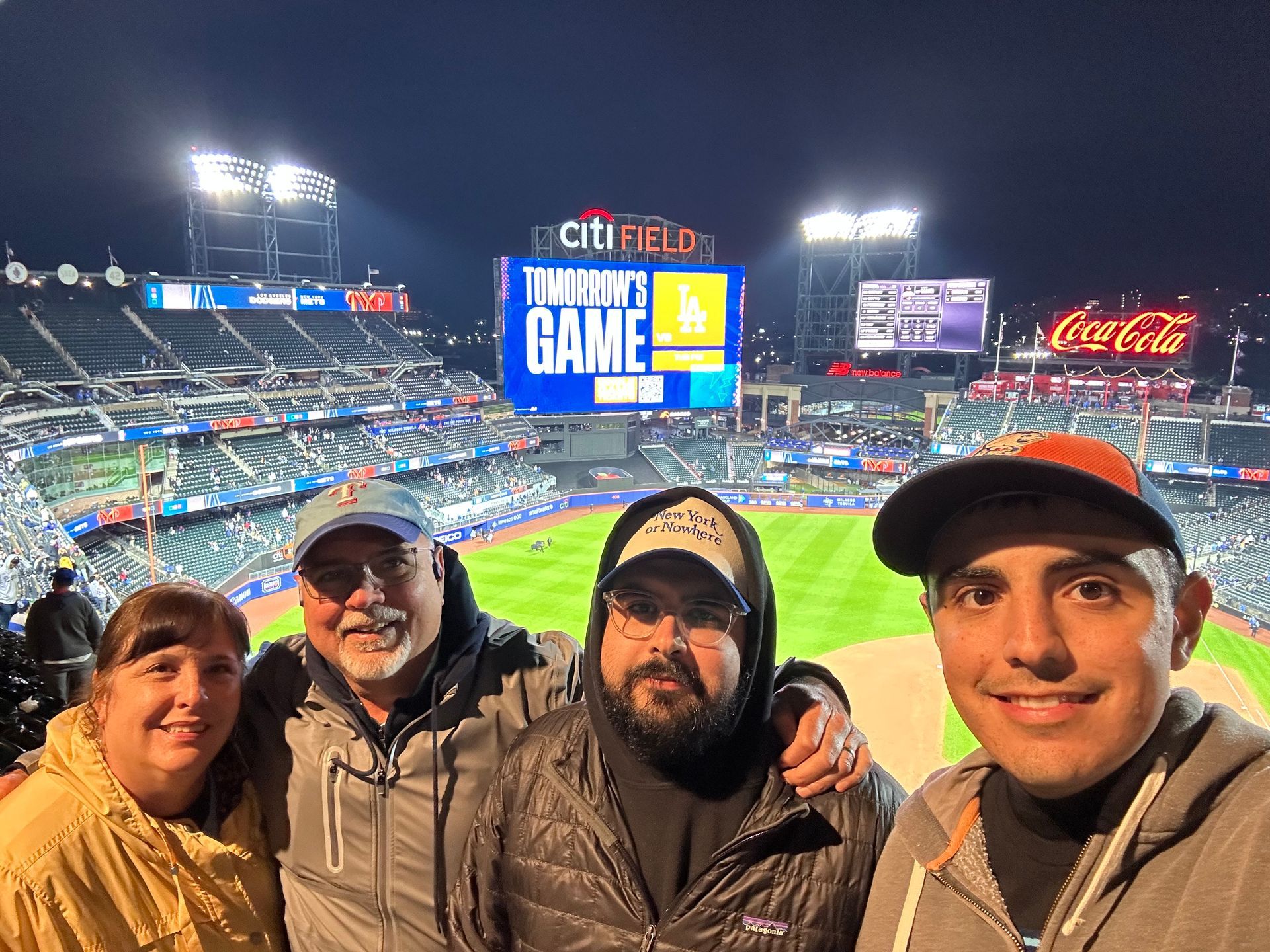 Four people smiling in a selfie at a night baseball game at Citi Field, with the brightly lit stadium in the background.