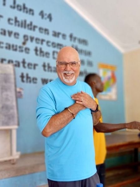 Smiling man in blue shirt clasping hands indoors, with another person in yellow in the background