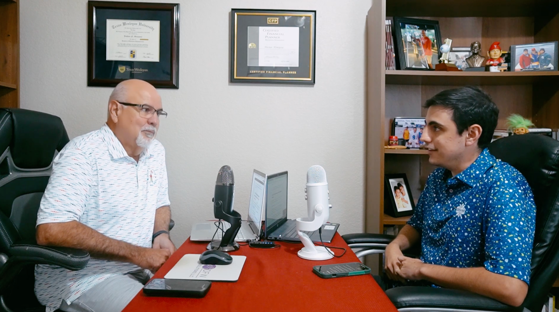 Two people sit at a red desk with microphones and a laptop between them, engaged in a conversation in an office setting.