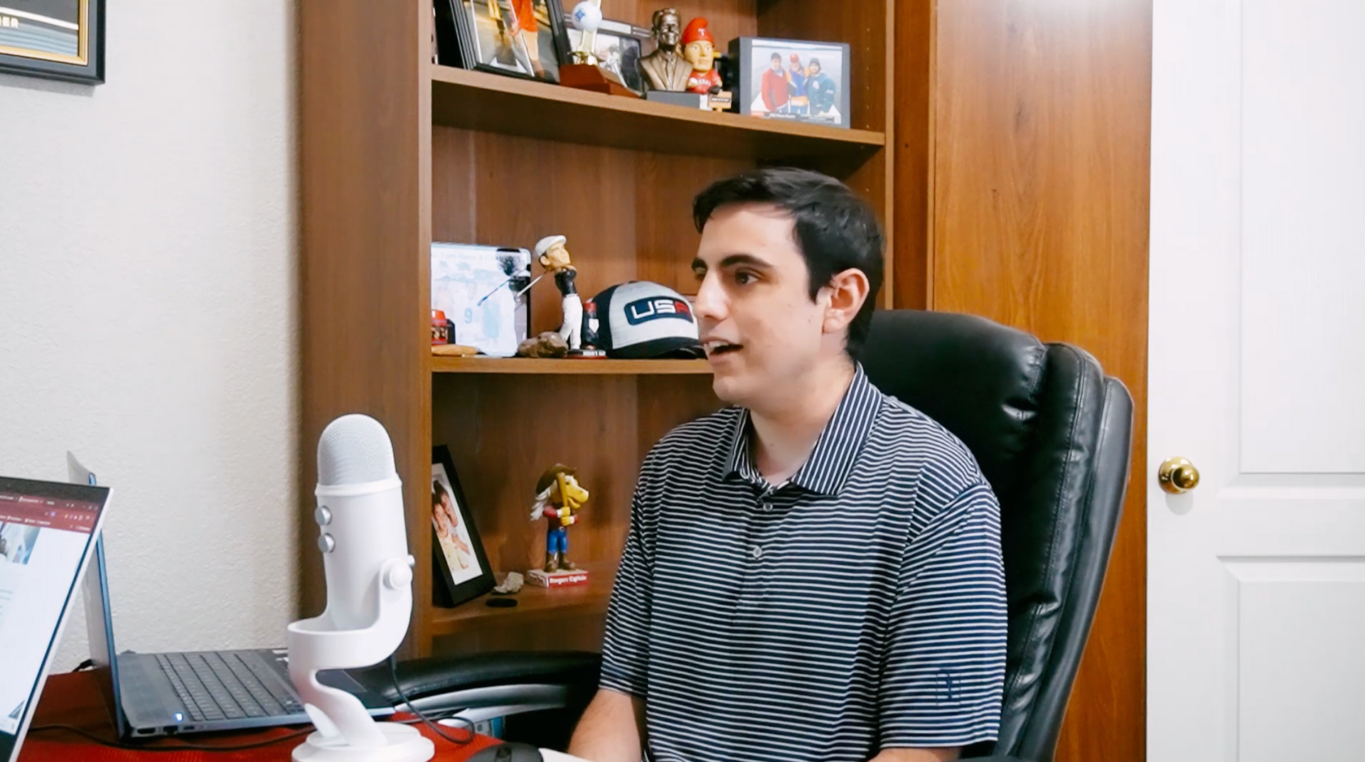 A person in a striped polo shirt sitting at a desk with a white microphone, in front of a wooden bookshelf.