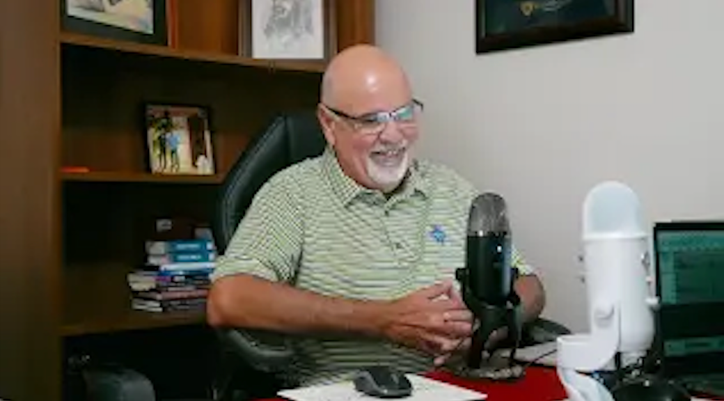 A person with glasses and a beard smiles while sitting at a desk with two microphones and a laptop in an office.