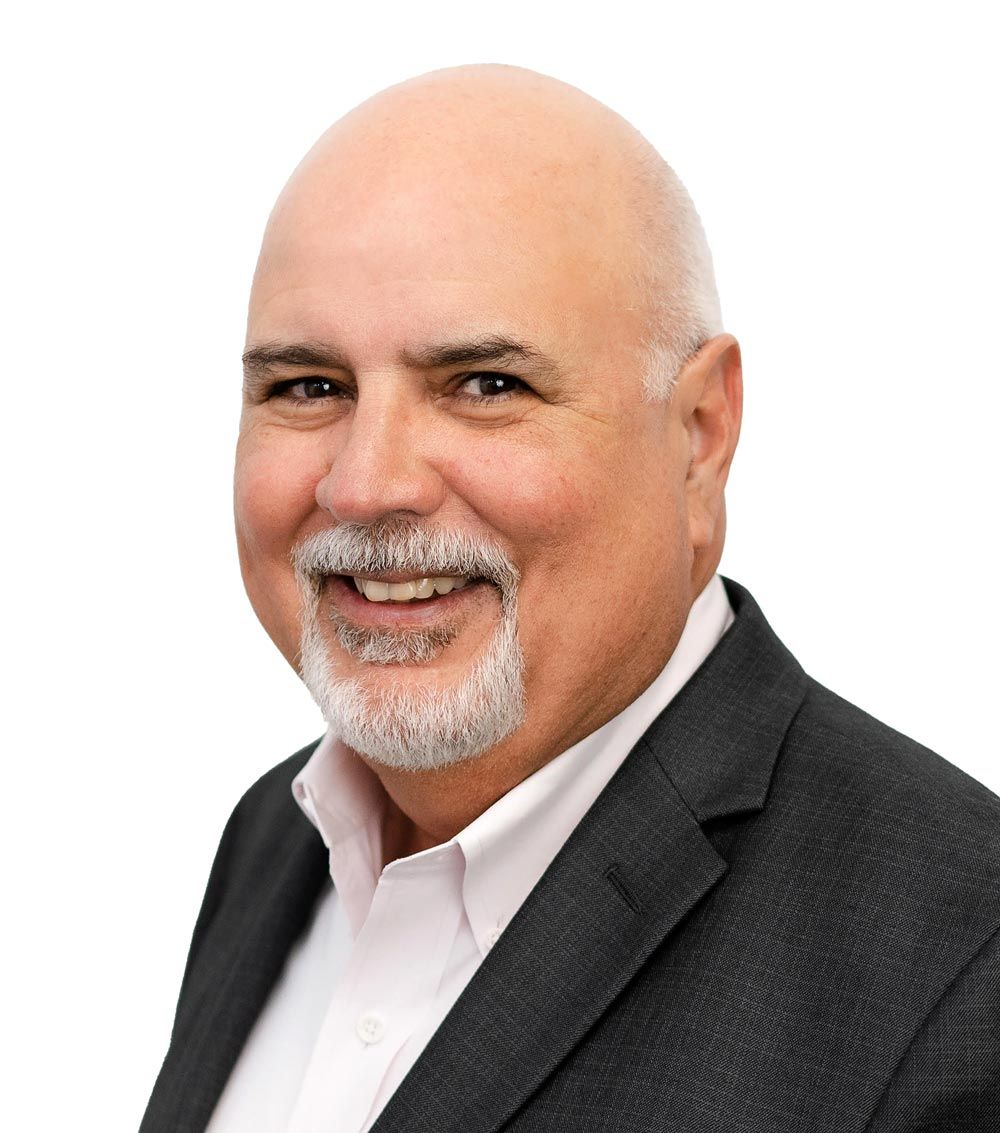 A person with a shaved head and facial hair, smiling, wearing a dark blazer over a white collared shirt against a white background.