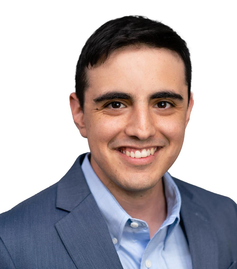 A person with short dark hair smiling, wearing a blue blazer over a light blue button-down shirt against a white background.
