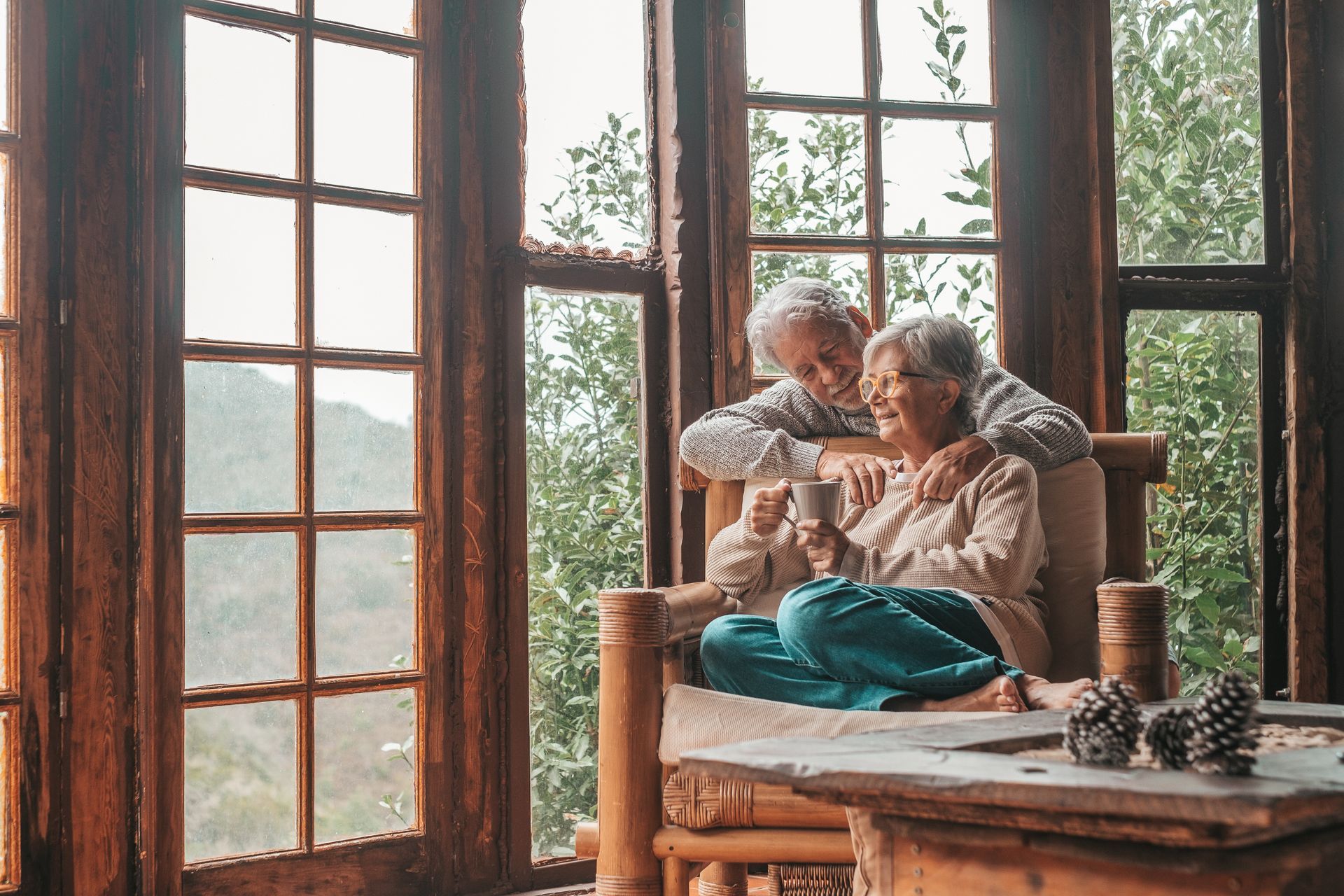 Two people sit on a wooden chair by large windows, looking at a photo album in a cozy room.