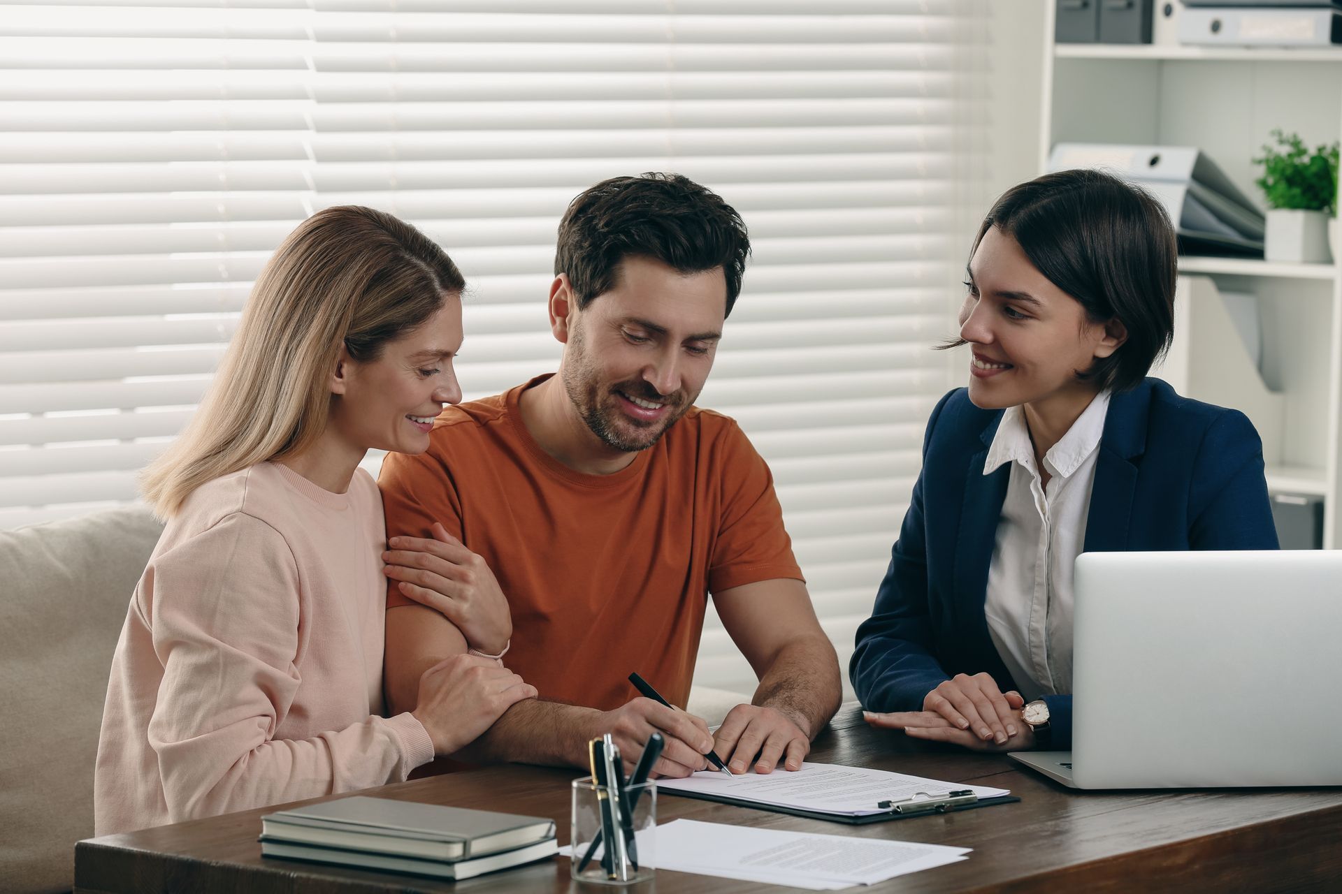 Three people discussing documents at a table with a laptop in a bright office