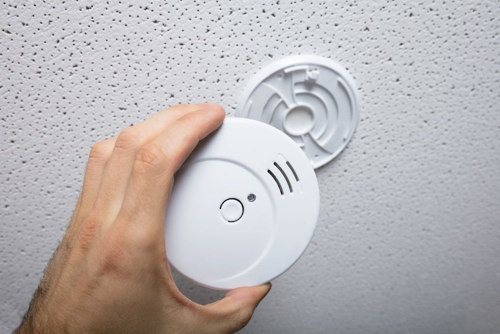 Hand Installing a White Smoke Detector on a Textured White Ceiling — FNQ Smoke Alarms & Maintenance in Innisfail, QLD