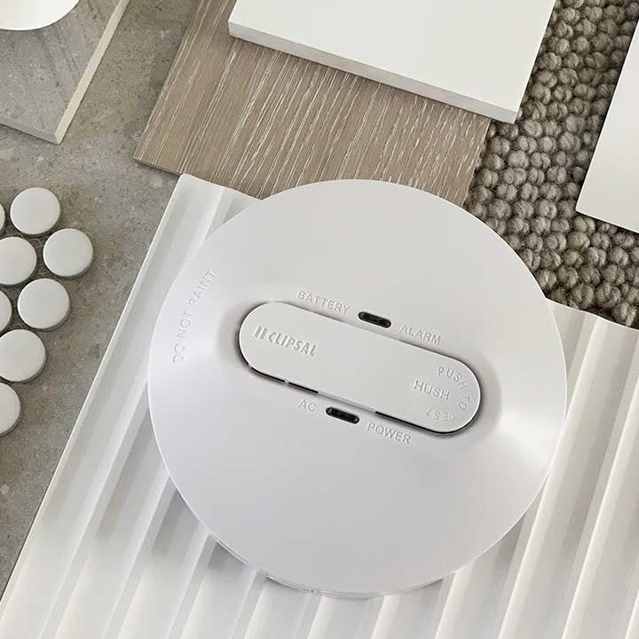 White Smoke Detector on a Textured Surface, With Tiles and Wood Samples in the Background — FNQ Smok