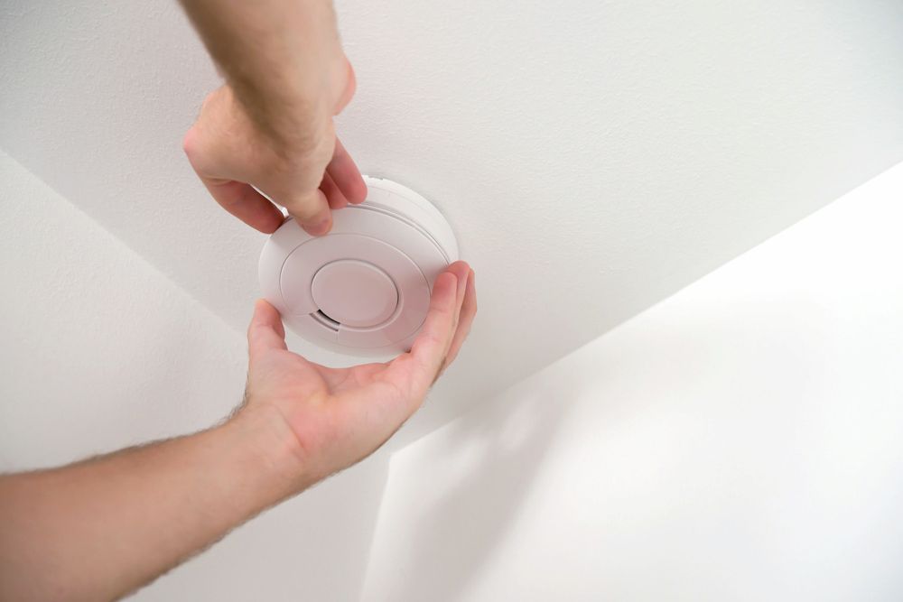 Hands Installing a White Smoke Detector on a White Ceiling — FNQ Smoke Alarms & Maintenance in Innisfail, QLD
