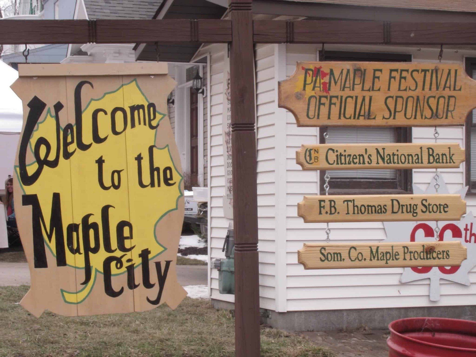 Sign welcoming to Maple City at a festival with official sponsor names.