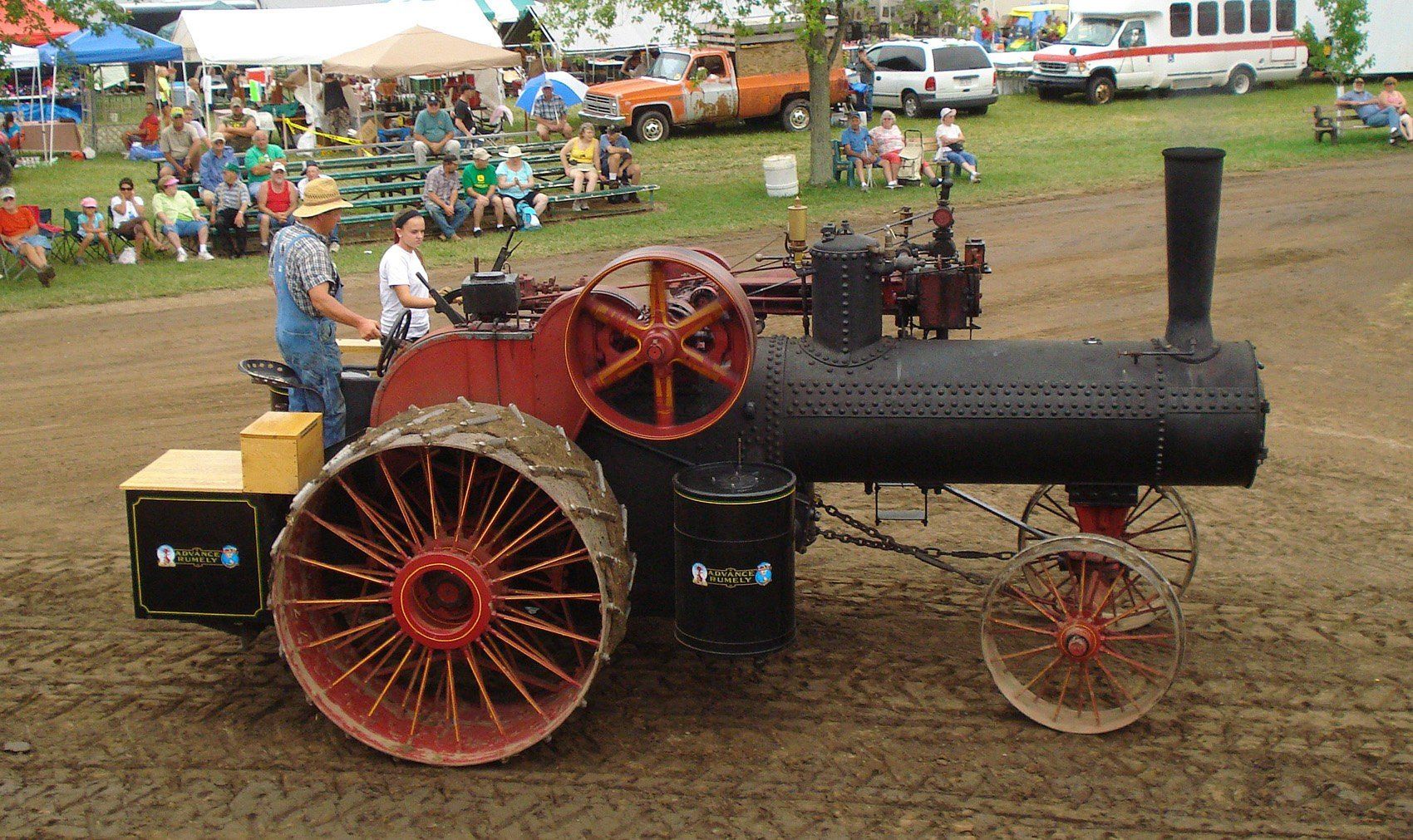 Steam engine tractor in a field with operators. Spectators watch in the background.