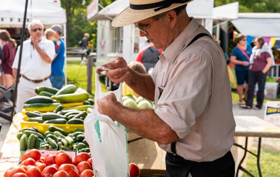 Man in straw hat at a farmer's market putting produce into a bag. Cucumbers and tomatoes on the table.