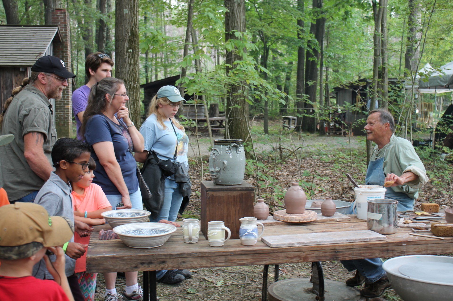 Potter demonstrating pottery to a group outdoors. People watch as he works at a table with clay and ceramic pieces.