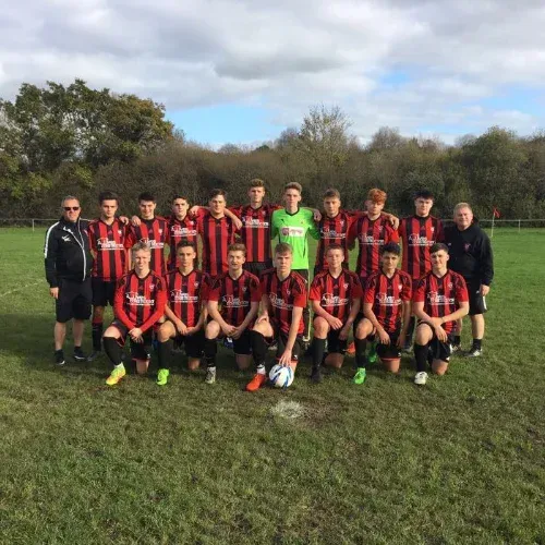 Football team in red and black striped kits posing together on a grass football pitch with their coaches.
