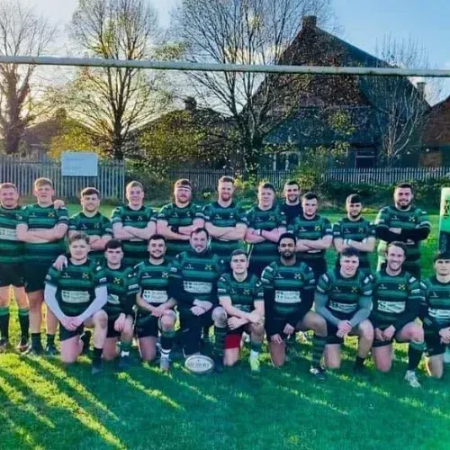 Rugby team posing on a field in green-and-black jerseys, with a goalpost and houses in the background