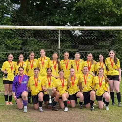 Women’s football team in yellow kits wearing medals and posing with a trophy in front of a goal.