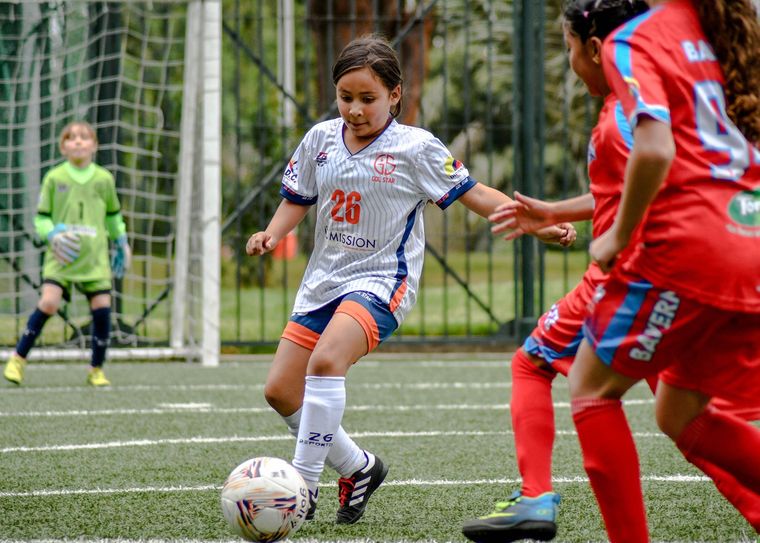 Youth soccer player in white dribbles the ball past red defenders on a field, with a goalkeeper in the goal.
