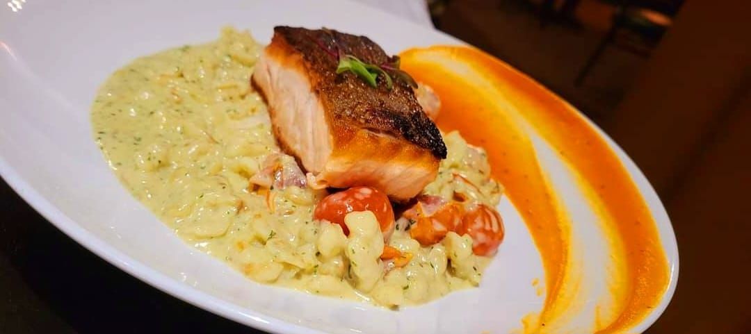 A seared salmon fillet served atop a bed of creamy risotto with tomatoes, garnished with a vibrant orange sauce swoosh.