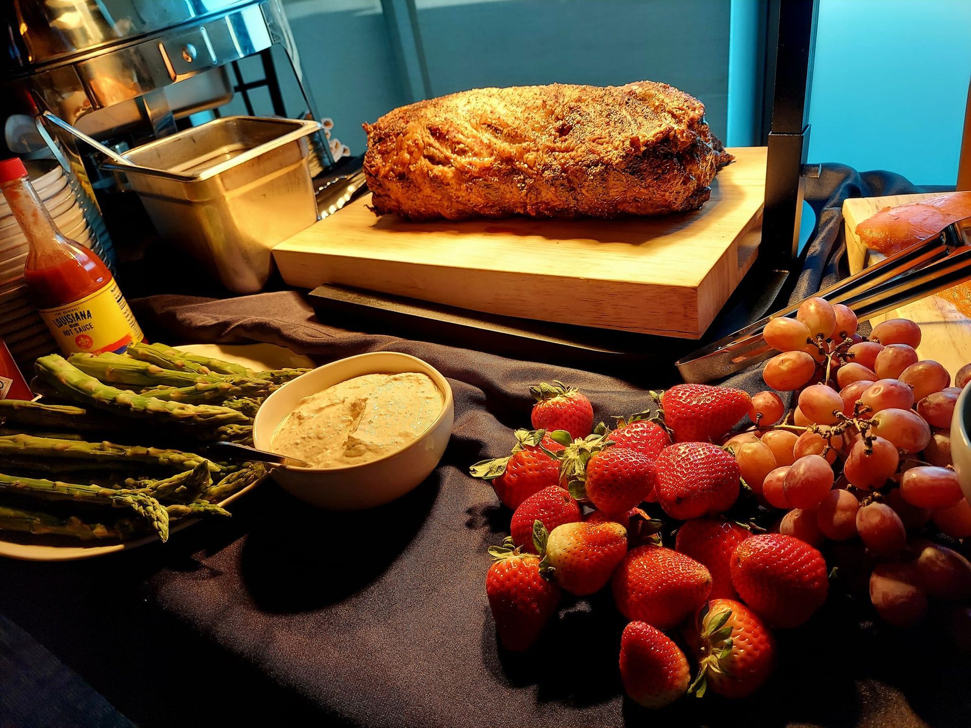 A buffet display featuring a roasted meat loaf, asparagus, a bowl of dip, fresh strawberries, and red grapes.