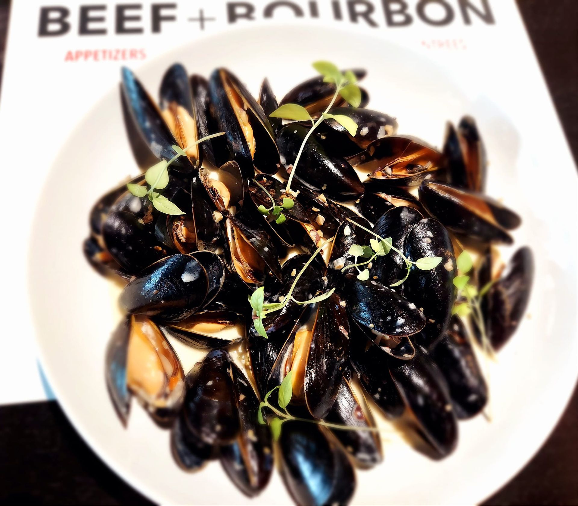 A white bowl filled with steamed black mussels, garnished with fresh herbs, resting on a menu titled Beef + Bourbon.