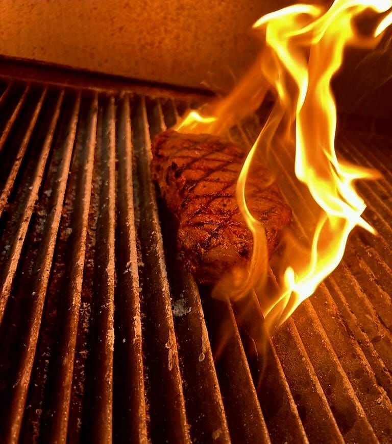 A thick steak with charred grill marks sears on a hot grate while flames lick up the side.