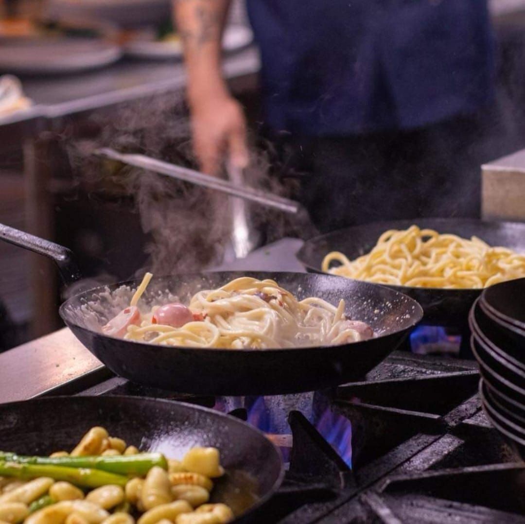 A chef cooks pasta and gnocchi in pans over open flames on a restaurant stove.