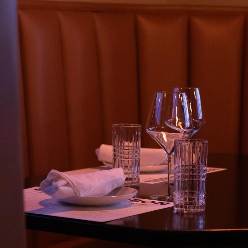 A restaurant table setting with two water glasses, wine glasses, and folded napkins on white linens against a booth.