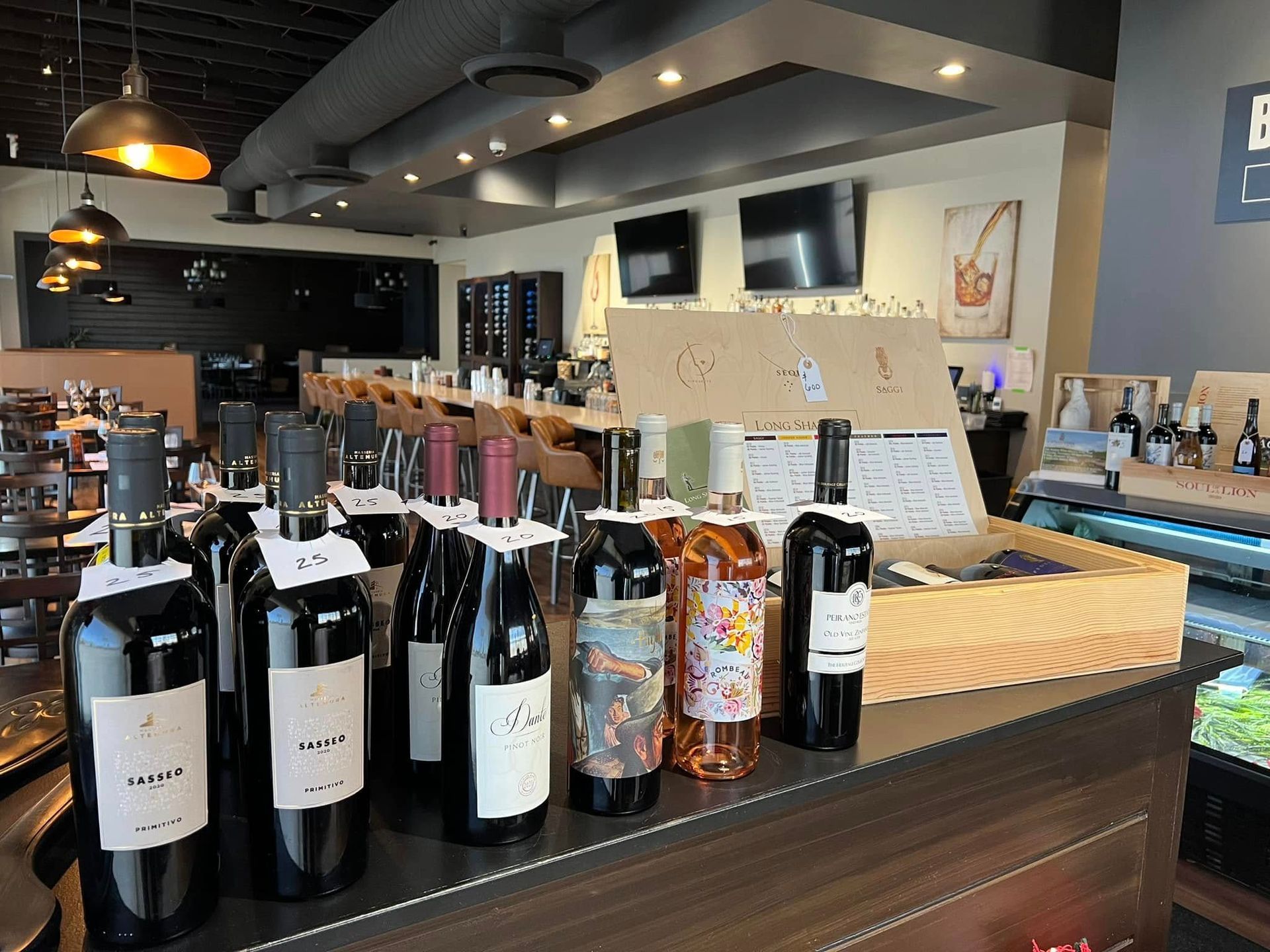 A row of wine bottles with descriptive tags sits on a counter in a dimly lit, modern bar with tables and TVs in the back.