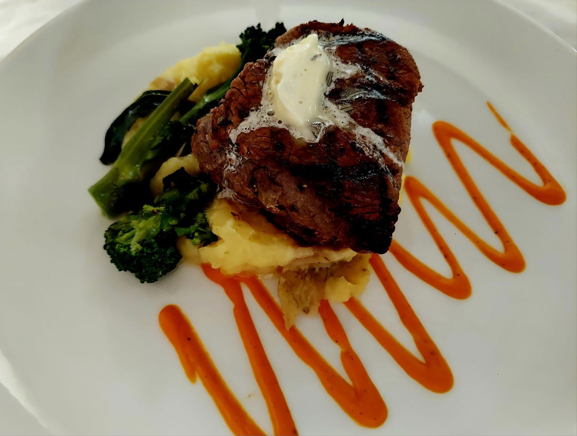 A grilled steak topped with melting butter, served with mashed potatoes and broccoli, garnished with orange sauce swipes.