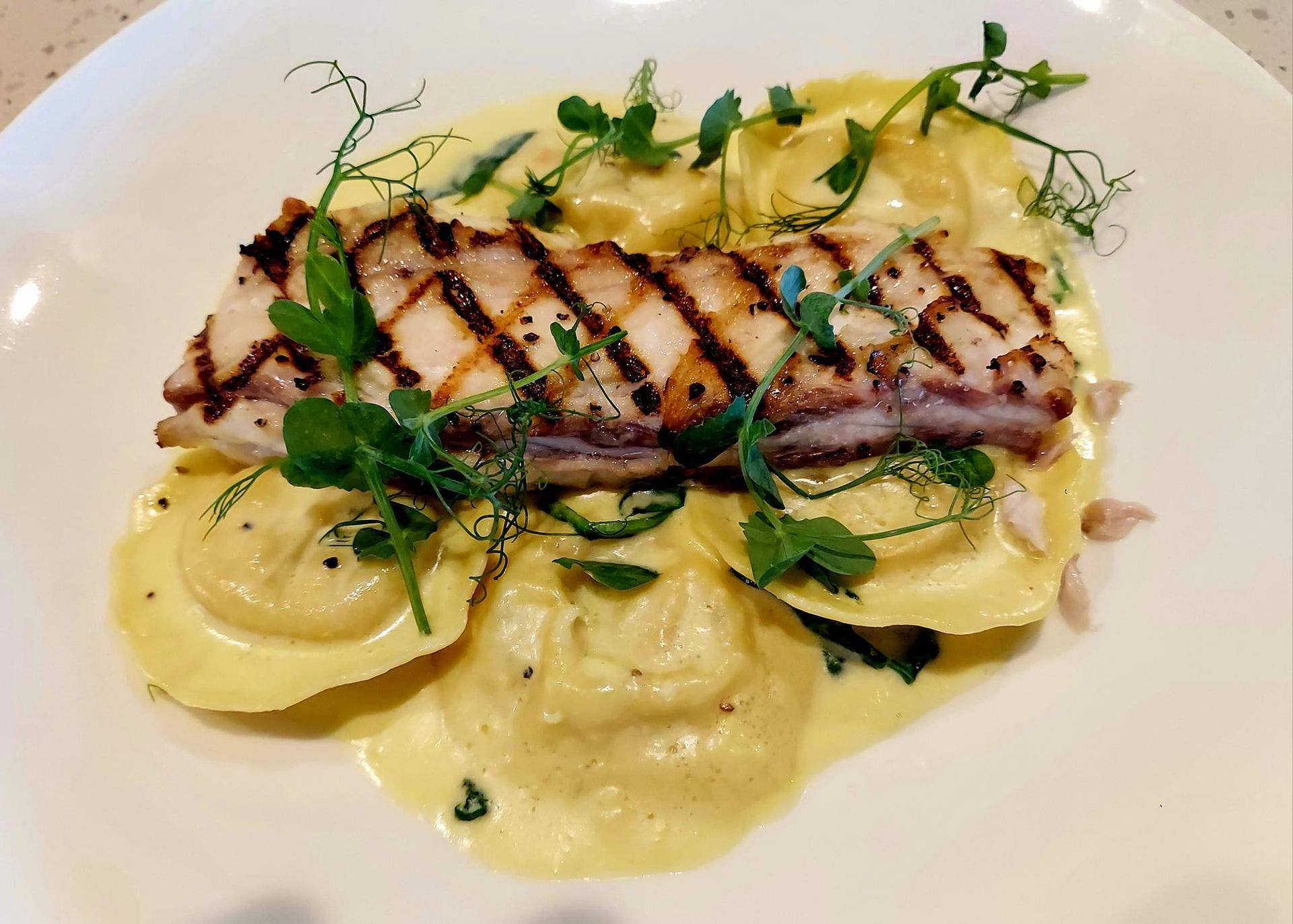 A grilled fish fillet served over ravioli in a creamy sauce, garnished with fresh pea shoots on a white plate.