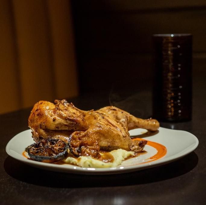 Roasted chicken served on a white plate with mashed potatoes, charred lemon, and sauce, beside a dark tumbler.
