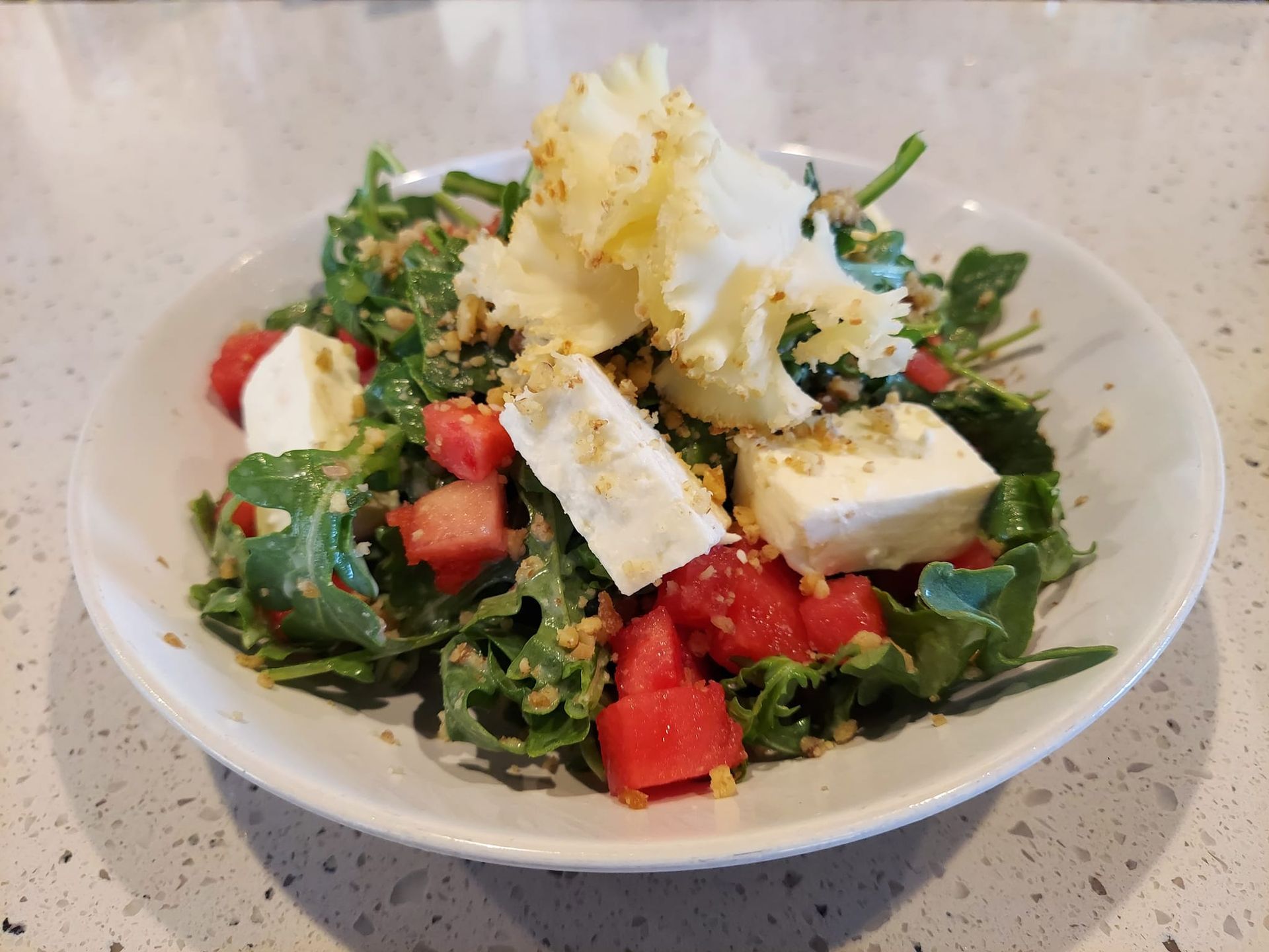 A salad of arugula, watermelon cubes, and feta cheese, topped with shaved cheese and nuts, served in a white bowl.