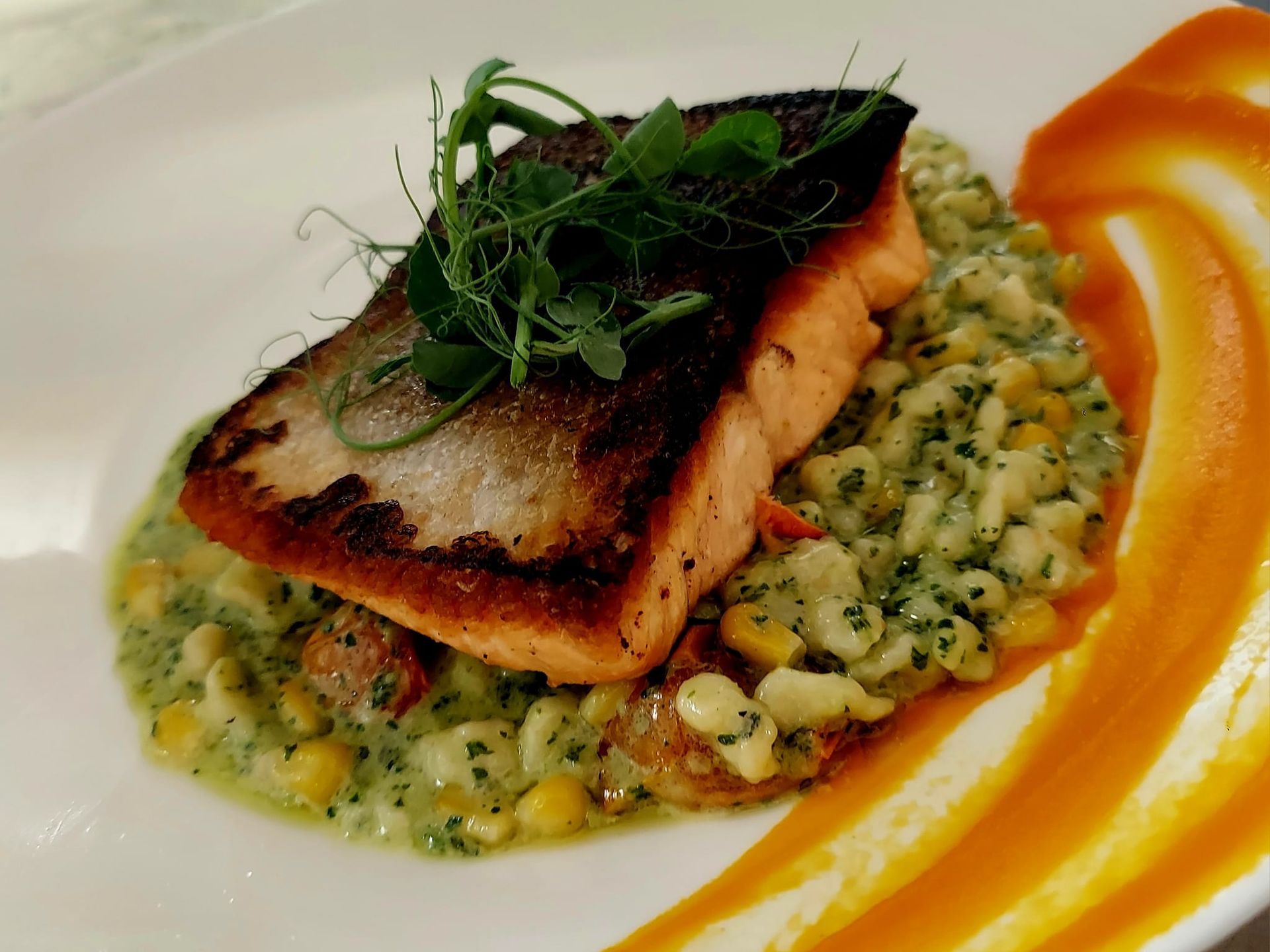 A seared salmon fillet atop a bed of creamy corn and herb succotash, garnished with microgreens and a vibrant orange puree.