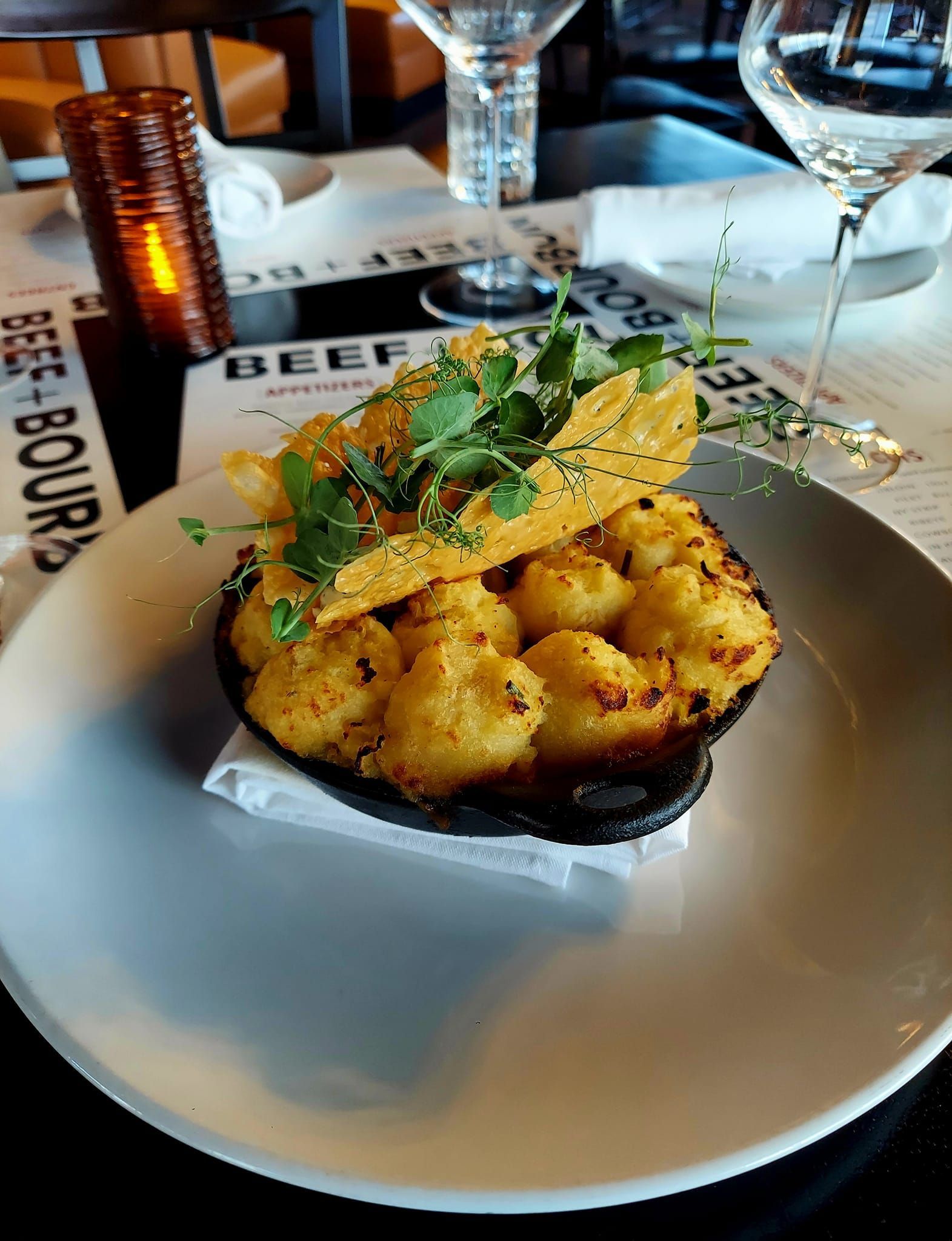 A small cast-iron skillet of tater tots topped with a large cheese crisp and herbs on a plate in a restaurant setting.