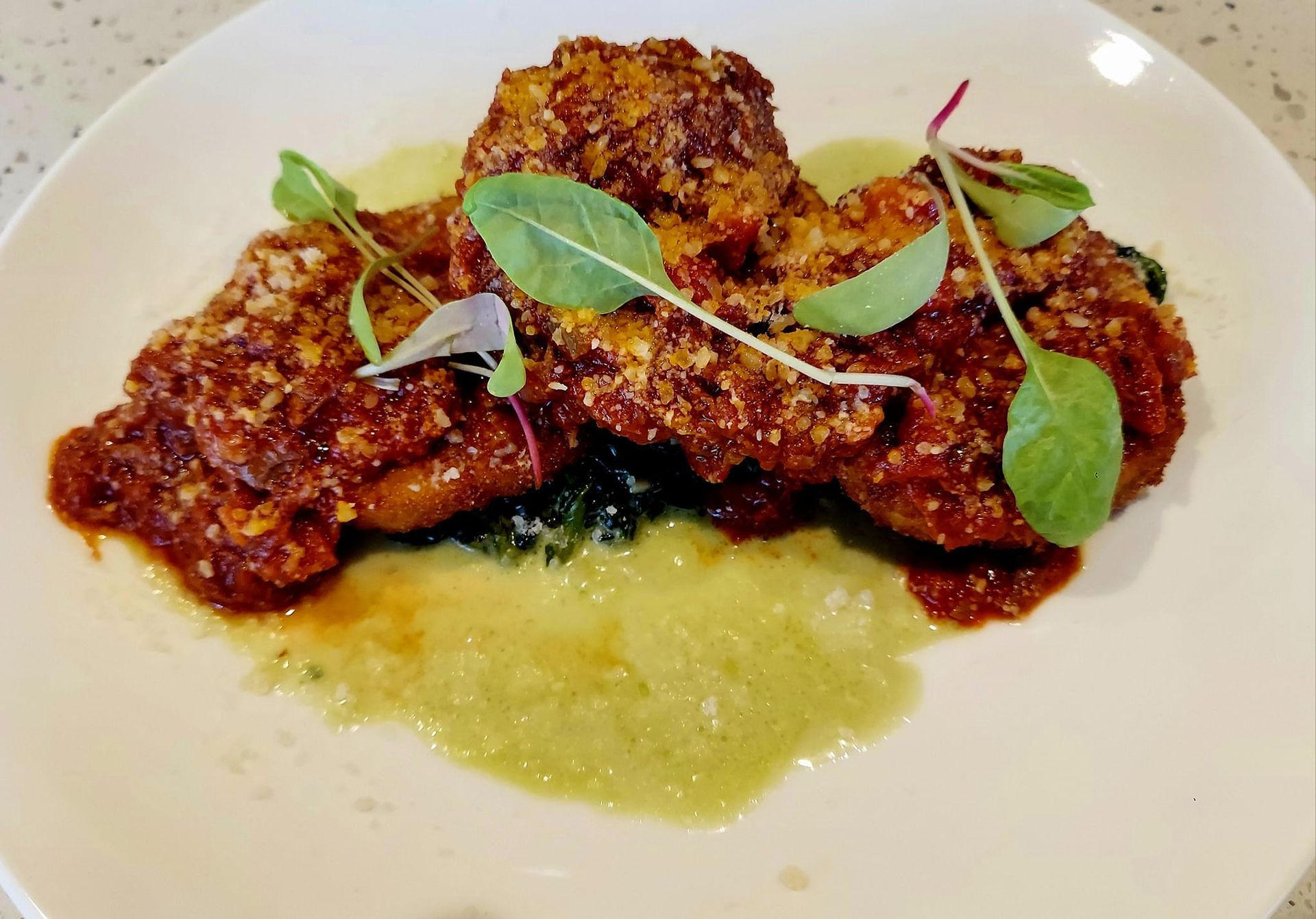Fried, breaded, and sauced patties served on a green sauce base, garnished with fresh greens on a white plate.