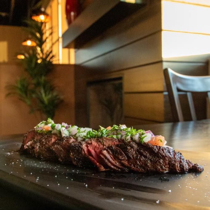 Sliced grilled steak topped with fresh pico de gallo served on a dark slate board in a restaurant setting.