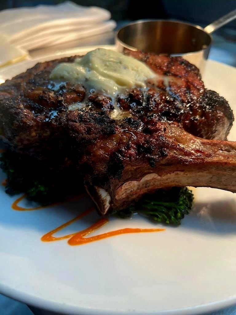 A seared bone-in steak topped with melting herb butter, served over greens on a white plate with sauce drizzles.