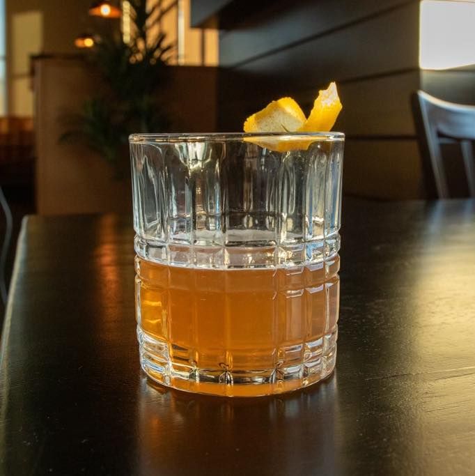 An amber-colored cocktail in a textured glass with a lemon peel garnish on a dark wooden table in a dimly lit restaurant.