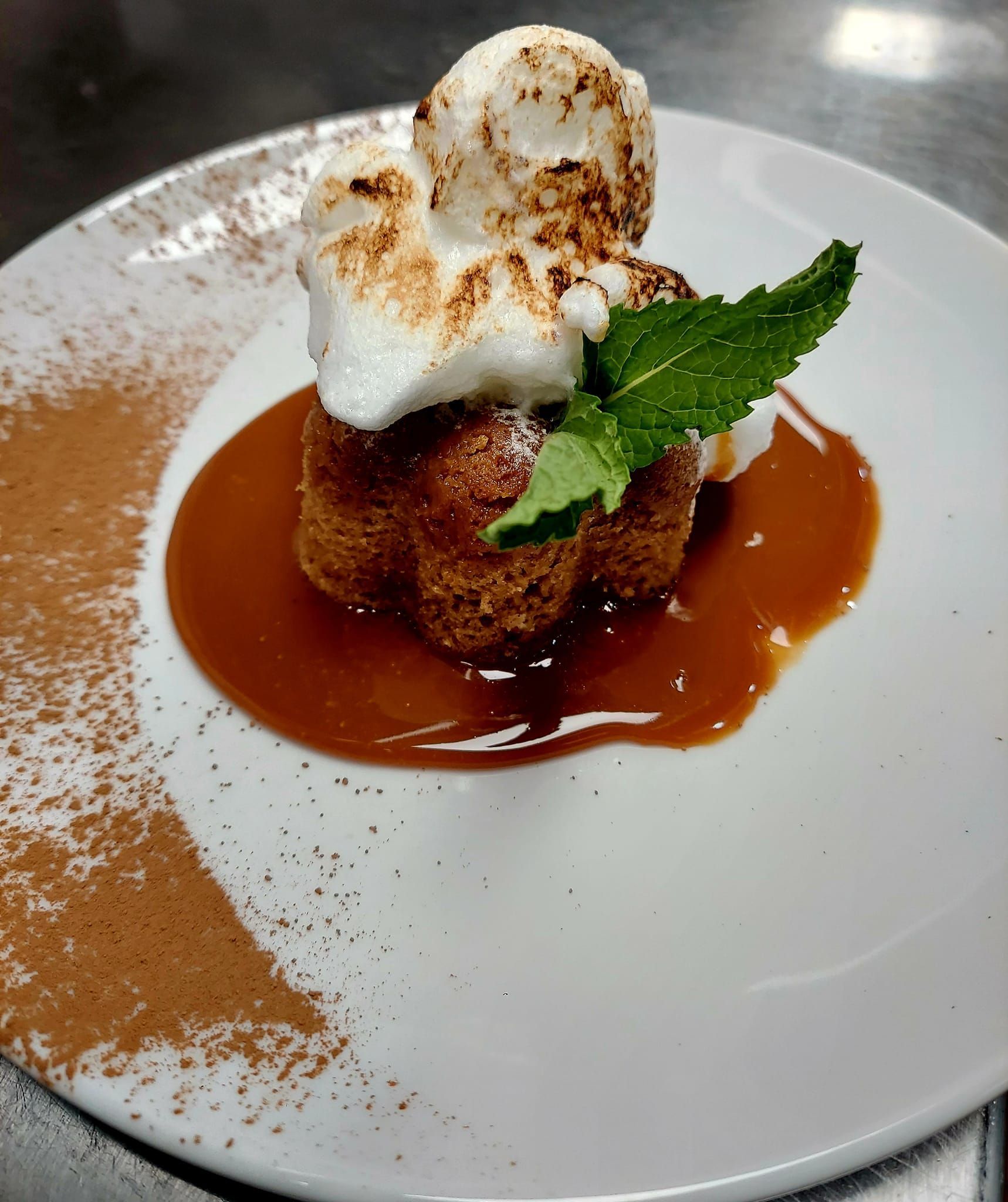 A small, browned cake topped with toasted meringue and a mint sprig, served in caramel sauce with a cocoa powder dusting.